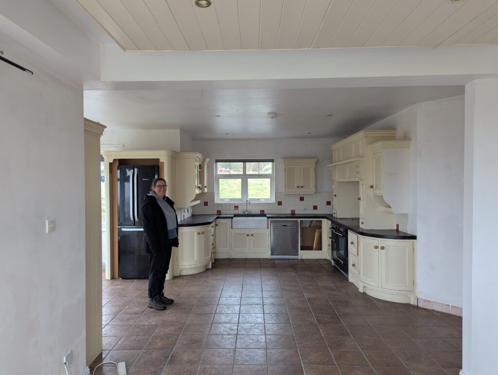View of a cream-painted wooden hand-built kitchen, terracotta tiled floor, woman in outdoor clothing standing to the left smiles at the camera