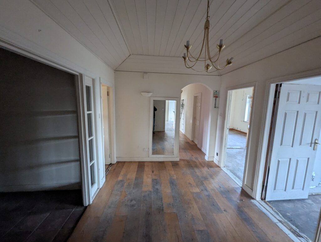 hallway of a bungalow, open doors to the right give glimpses of rooms beyond. The wooden floor is patchy and varied in colour